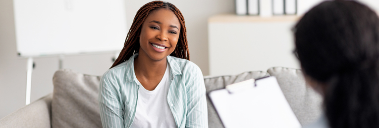 A person smiling is seated across from another individual (whose face is not visible) who is holding a pad with paper, as if taking notes or conducting an interview.